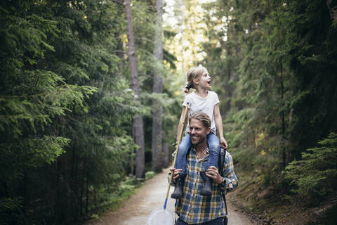 Lächelnder Vater trägt seine Tochter auf der Schulter beim Spaziergang im Wald, lizenzfreies Stockfoto