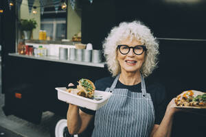 Portrait of senior owner with street food standing against commercial land vehicle in city - MASF20241