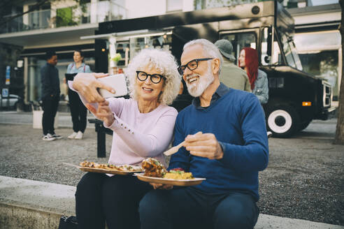 Lächelnde ältere Frau, die ein Selfie mit einem Mann macht, der eine Mahlzeit gegen einen Essenswagen in der Stadt isst - MASF20247