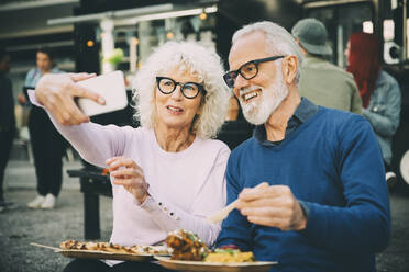 Senior woman taking selfie with smiling man eating meal against food truck in city - MASF20248