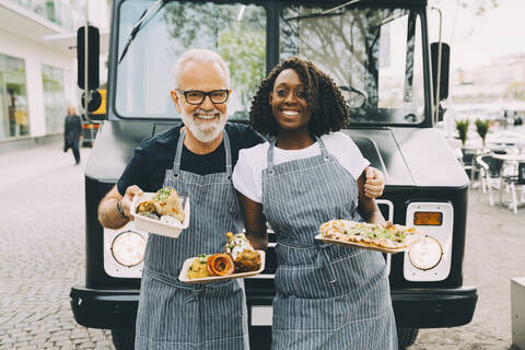 Smiling senior owner with assistant holding food plate while standing against commercial land vehicle stock photo