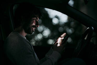 Smiling man using mobile phone while sitting in car at night - EBBF01018