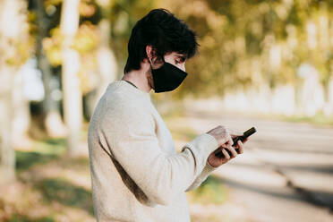 Mann mit Gesichtsmaske, der ein Mobiltelefon benutzt, während er im Wald steht - EBBF01021