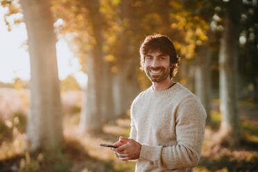 Smiling man using mobile phone while standing at forest - EBBF01031