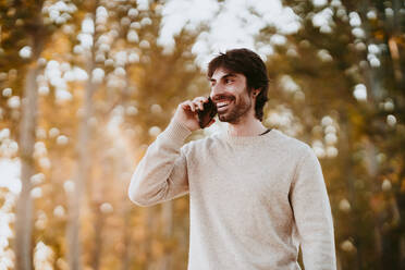 Lächelnder Mann, der wegschaut, während er im Wald mit seinem Handy telefoniert - EBBF01036