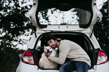 Mann umarmt Hund, während er im Kofferraum eines Autos im Wald sitzt - EBBF01044