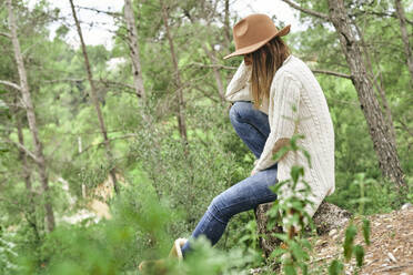 Woman wearing hat while sitting in forest - VEGF02998