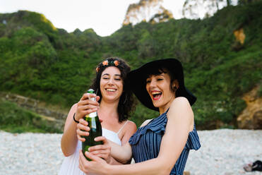 Fröhliches lesbisches Paar, das sich am Wochenende am Strand entspannt und eine Glasflasche mit einem alkoholischen Getränk öffnet - ADSF16951