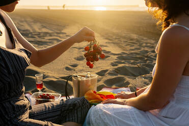 Seitenansicht einer zufriedenen Frau, die ihre Freundin mit Trauben füttert, während sie sich am Meeresufer bei Sonnenuntergang im Sommer im Gegenlicht entspannt - ADSF16956