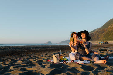 Couple of lesbians resting on seashore with alcohol and taking photo on smartphone while spending vacation together - ADSF16957