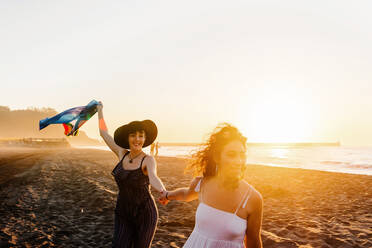 Glückliche Frauen, die eine mehrfarbige Flagge am Strand hissen - ADSF16962