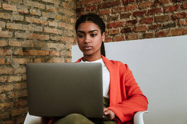 Focused elegant African American woman sitting with laptop in armchair against brick wall in loft apartment - ADSF17013