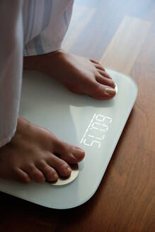 Draufsicht auf eine gesichtslose, barfuß gehende Frau im gemütlichen Pyjama, die auf einer digitalen Gewichts- und Körperfettwaage steht, deren Display ein gesundes Gewicht von 60 kg auf dem Badezimmerboden am Morgen anzeigt - ADSF17015