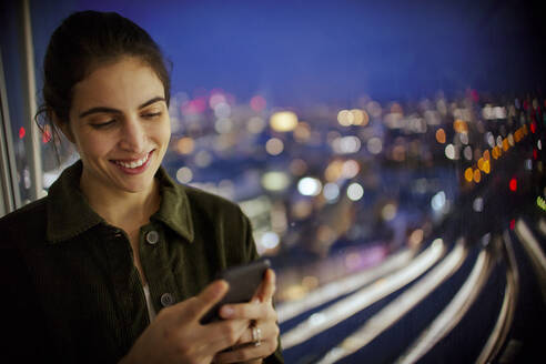Lächelnde Geschäftsfrau mit Smartphone, die bis spät in die Nacht am Bürofenster arbeitet - CAIF29712