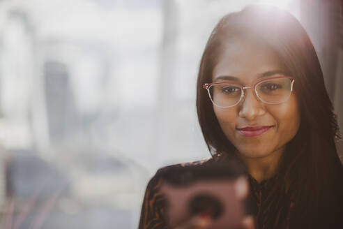 Lächelnde Geschäftsfrau mit Smartphone am sonnigen Fenster - CAIF29818