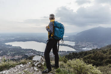 Mature man with backpack looking at lake and city against sky during sunset - MCVF00633