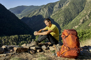 Mann schaut weg, während er auf einem Stein sitzt und eine Kaffeetasse in einem Wald an einem sonnigen Tag hält - VEGF03021