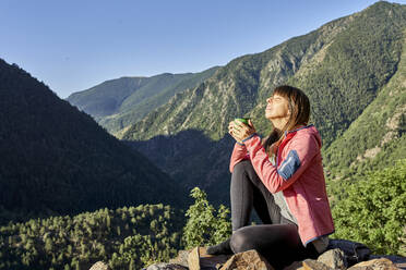 Mittlere erwachsene Frau, die eine Kaffeetasse in der Hand hält, während sie auf einem Stein im Wald sitzt, an einem sonnigen Tag - VEGF03029