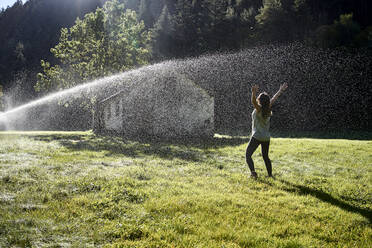 Woman standing in front of sprinkler on grass during sunny day - VEGF03031