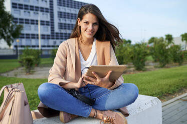Lächelnde junge Frau mit digitalem Tablet auf einer Bank in einem öffentlichen Park sitzend - KIJF03353