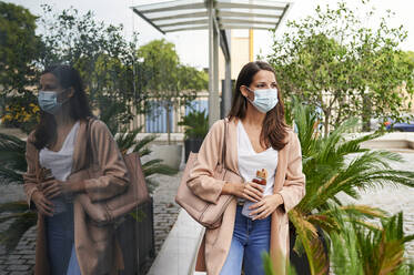 Young woman wearing protective face mask holding reusable bottle standing on street - KIJF03359