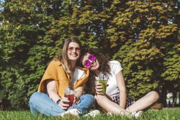 Lächelnde Freundinnen sitzen mit frischer Limonade im Gras im Park an einem sonnigen Tag - VYF00147