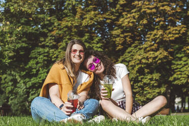 Smiling female friends sitting with fresh lemonade on grass at park during sunny day - VYF00147