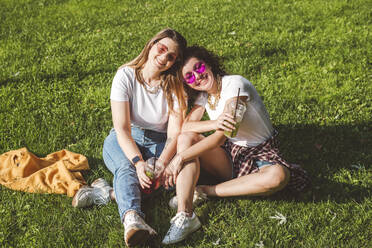 Glückliche Freundinnen sitzen mit Limonade im Gras im Park an einem sonnigen Tag - VYF00149
