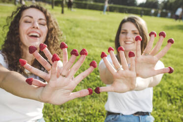 Playful female friends enjoying with fresh raspberries on fingers at park during sunny day - VYF00152