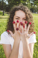 Verspielte junge Frau mit Himbeeren auf den Fingern im Park an einem sonnigen Tag - VYF00153