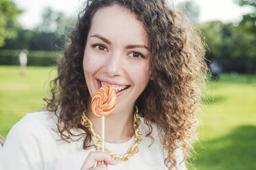 Schöne glückliche junge Frau mit lockigem braunem Haar isst Lutscher im Park - VYF00165