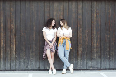 Beautiful women looking at each other while leaning against brown wooden wall - VYF00179