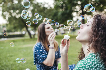 Beautiful female friends enjoying while blowing bubbles from wands at park - VYF00202