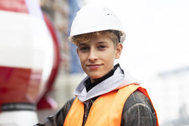 Selbstbewusster männlicher Jugendlicher mit Schutzhelm und reflektierender Kleidung auf einer Baustelle - SGF02702