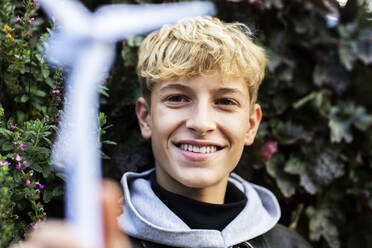 Lächelnder blonder Teenager, der ein Windradmodell gegen Pflanzen hält - SGF02703