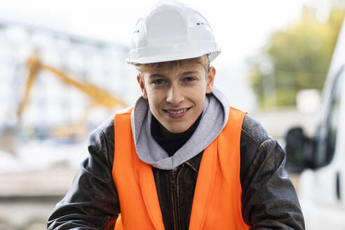 Lächelnder jugendlicher Bauauszubildender mit Schutzhelm und reflektierender Kleidung auf der Baustelle - SGF02705