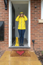 Woman wearing raincoat standing on doorway at home during rainy season - WPEF03470