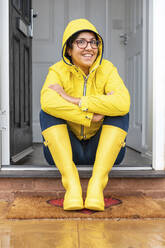 Smiling woman wearing raincoat while sitting on doorway at house during rainy season - WPEF03473