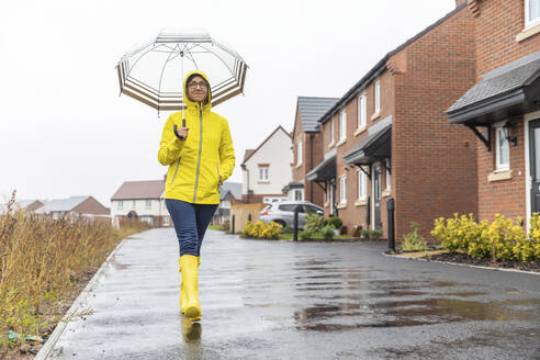 Frau mit Händen in den Taschen, die einen Regenschirm hält, wacht auf der Straße in der Stadt während der Regenzeit - WPEF03477