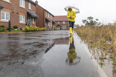 Woman holding umbrella walking in puddle on street during rainy season - WPEF03479