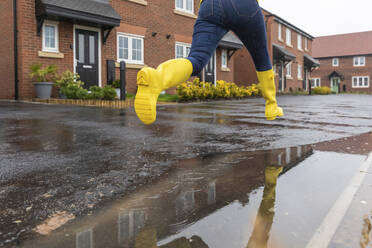 Close-up of woman jumping over water puddle on street during rainy season - WPEF03483