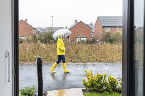 Mittlere erwachsene Frau mit Händen in den Taschen, die einen Regenschirm hält und auf der Straße wacht, während es regnet - WPEF03497