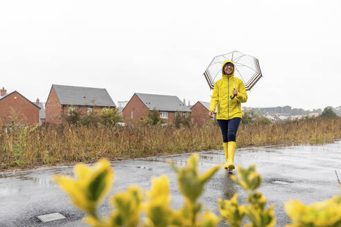 Lächelnde Frau mit Regenschirm, die wegschaut, während sie in der Regenzeit auf der Straße geht - WPEF03498