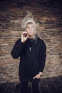 Junger Mann rauchend mit Händen in den Taschen an der Wand stehend - ACPF00854