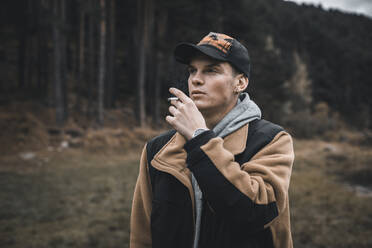 Young man looking away while smoking standing in forest during autumn - ACPF00857