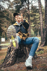 Junger Mann, der im Herbst auf einem Baumstamm im Wald sitzt und gestikuliert - ACPF00868