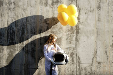Girl holding balloon while standing against wall on sunny day - GGGF00001