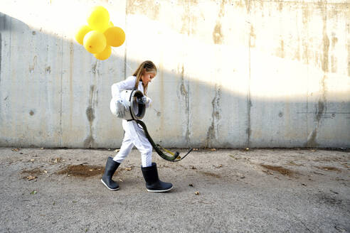 Little girl walking with space helmet and holding balloon on street during sunny day - GGGF00002