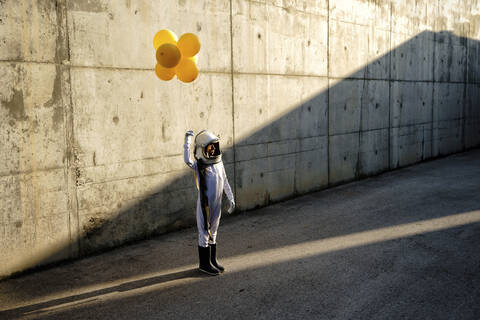 Little girl wearing space helmet holding balloon standing on street against wall stock photo