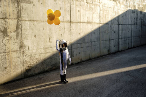 Little girl wearing space helmet holding balloon standing on street against wall - GGGF00004